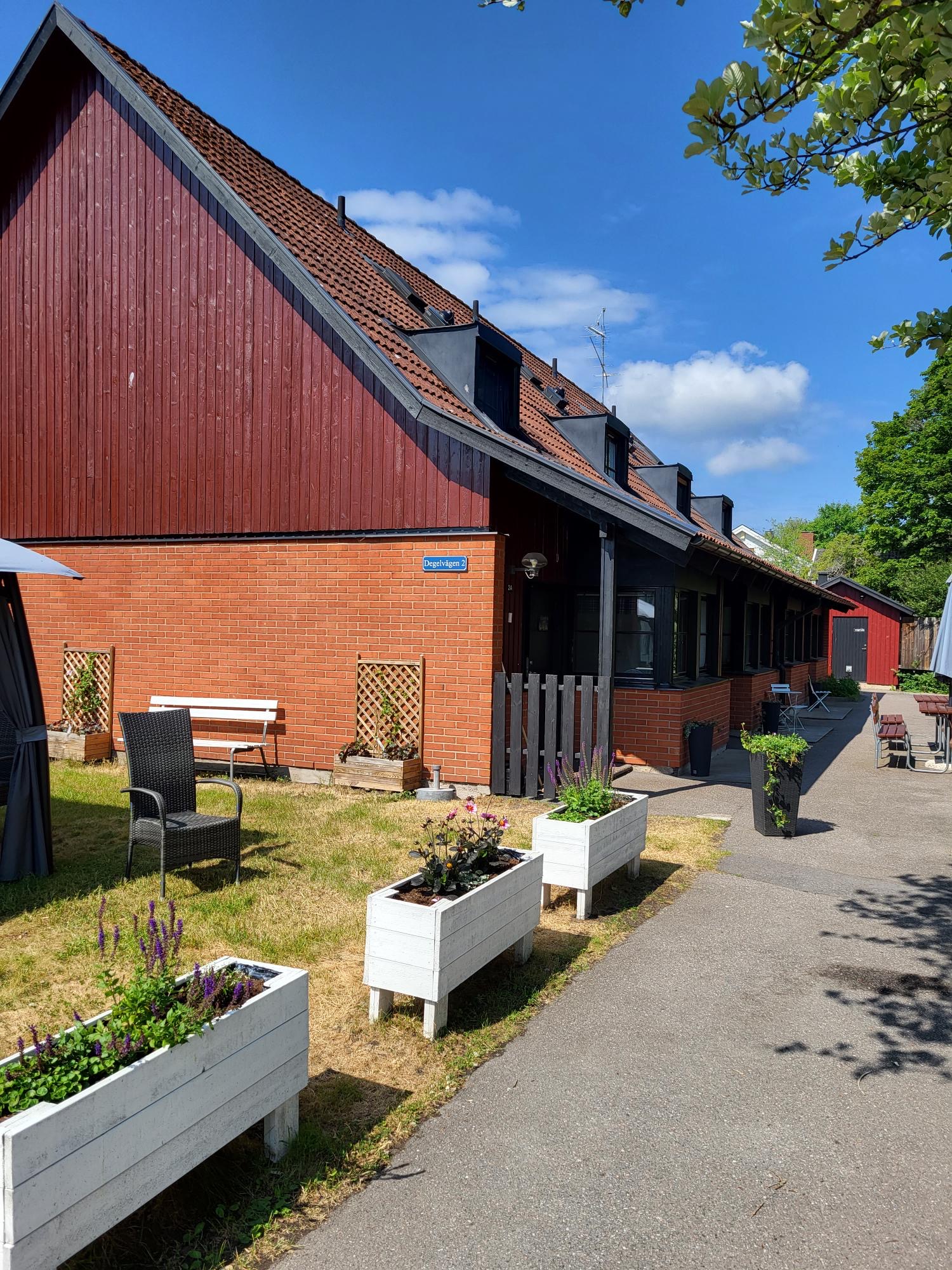 En röd tegel- och träbyggnad med branta tak, omgiven av gräsmatta, vita blomlådor och träd. Klarblå himmel i bakgrunden