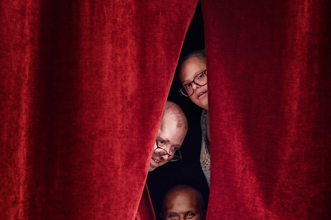 Tre deltagare på daglig verksamhet kikar fram i en röd teaterridås springa.