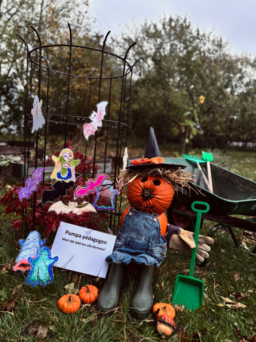 Pumpa med hatt, kropp i snickarbyxor och gummistövlar sitter utomhus omgiven av pysslade spöken och fjärilar. En skylt med text: "Pumpapedagogen - Med rätt stöd kan all blomstra!"