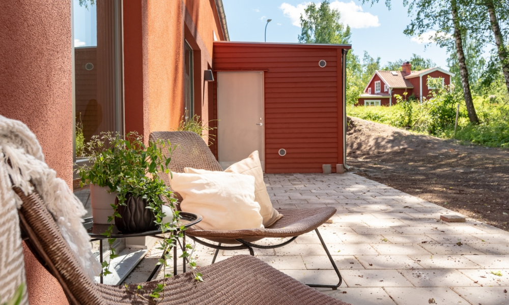 Avkopplande uteplats med moderna rottingmöbler och grön växt, belägen vid rött hus i lantlig miljö, solig sommardag